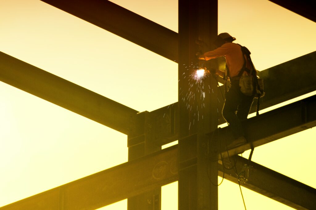 A worker welds together steel beams. The sky is a warm gold color.